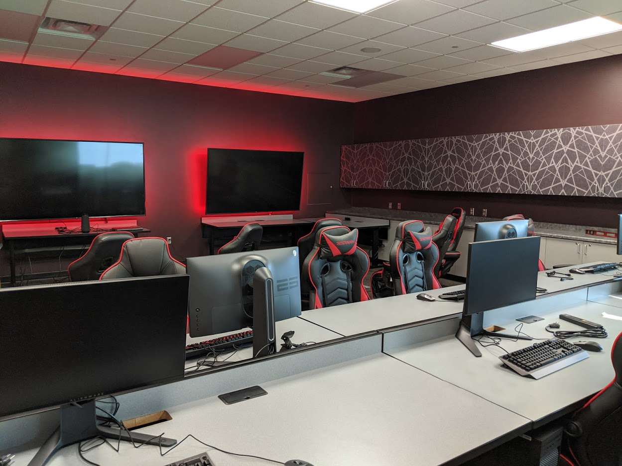 Two large monitors backlit with LED lighting, several computer stations with red and black chairs